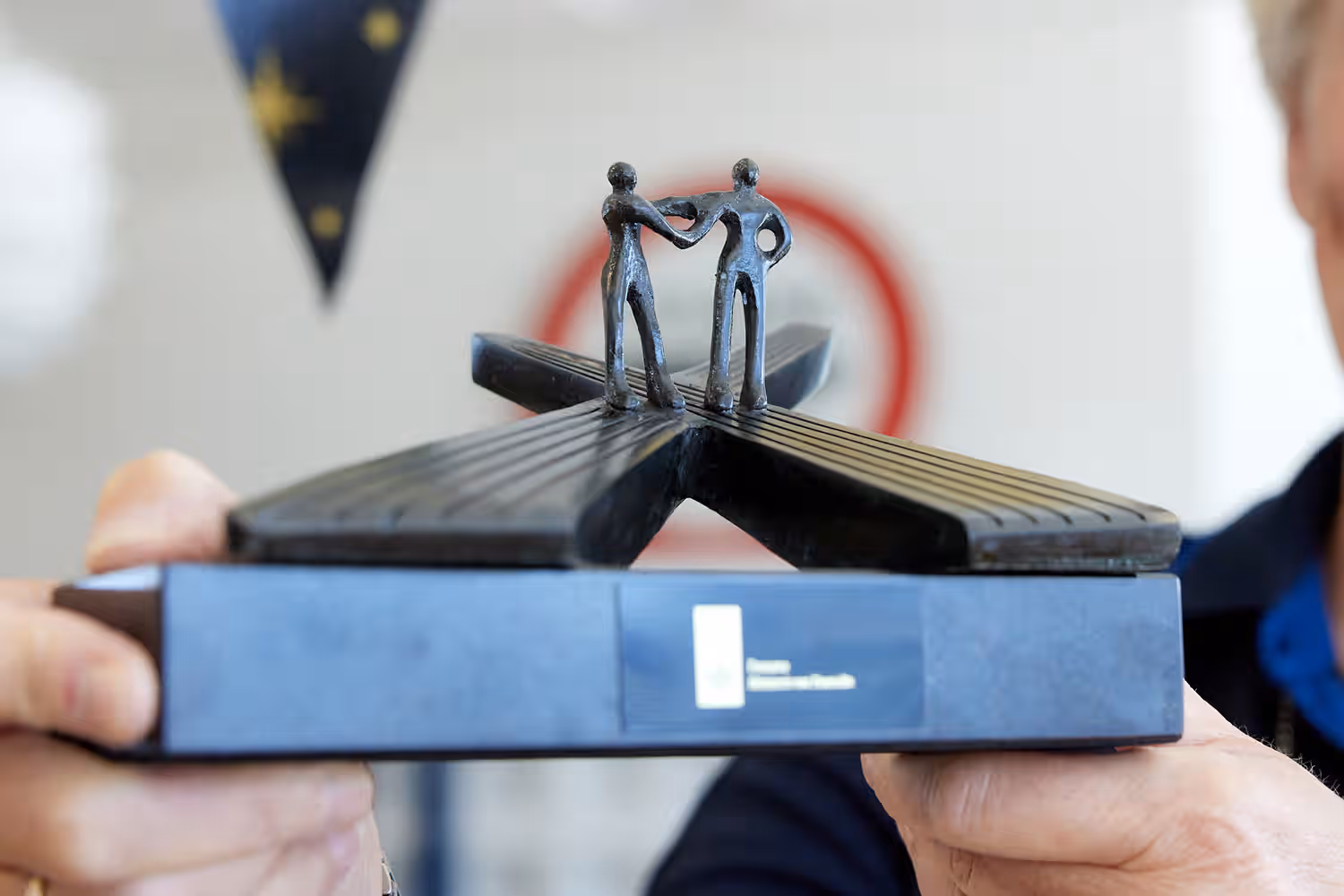 Afbeelding van een afscheidscadeau van de Doaune. Het is metalen beeldje met op de basis een klein logo van de Douane. Op de basis staat een structuur dat lijkt op wegen die elkaar kruisen. In het midden van het kruispunt staan twee menselijke figuren die elkaar de hand schudden.